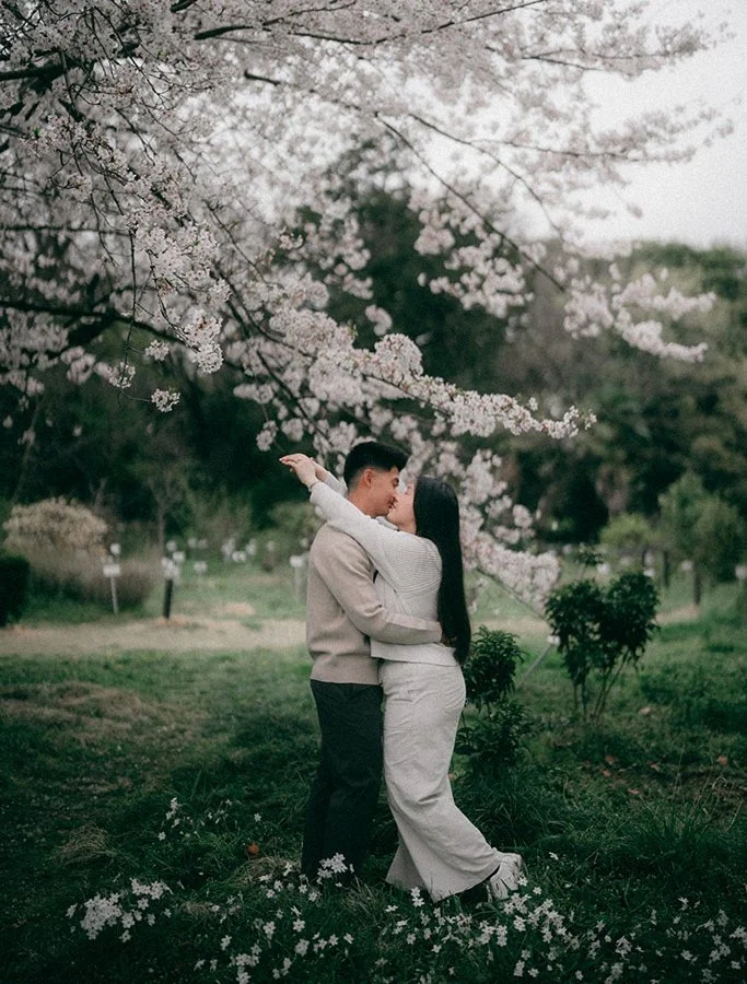 A couple is standing outdoors under a flowering tree, embracing and about to kiss. The woman has long black hair and is wearing a gray outfit, while the man has short dark hair and is wearing a beige jacket. They are surrounded by grass and small whi