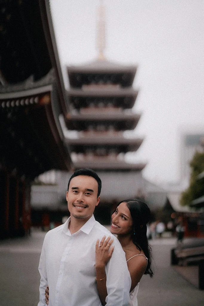 A happily smiling couple standing outdoors with a pagoda-style building in the background, lit up at dusk.