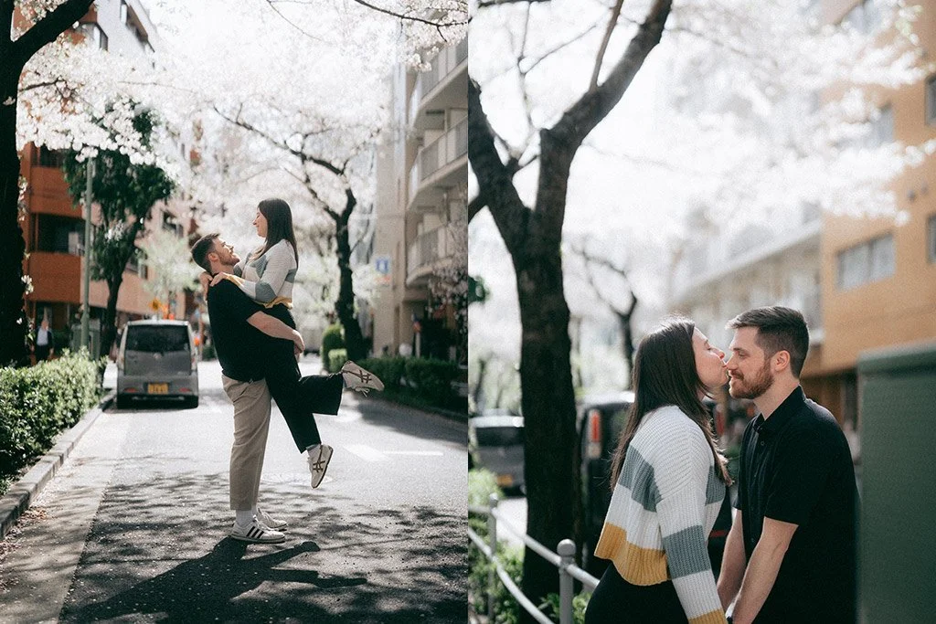 A couple in a city street with cherry blossoms, the man lifting the woman, both smiling, with cars and apartment buildings in the background.