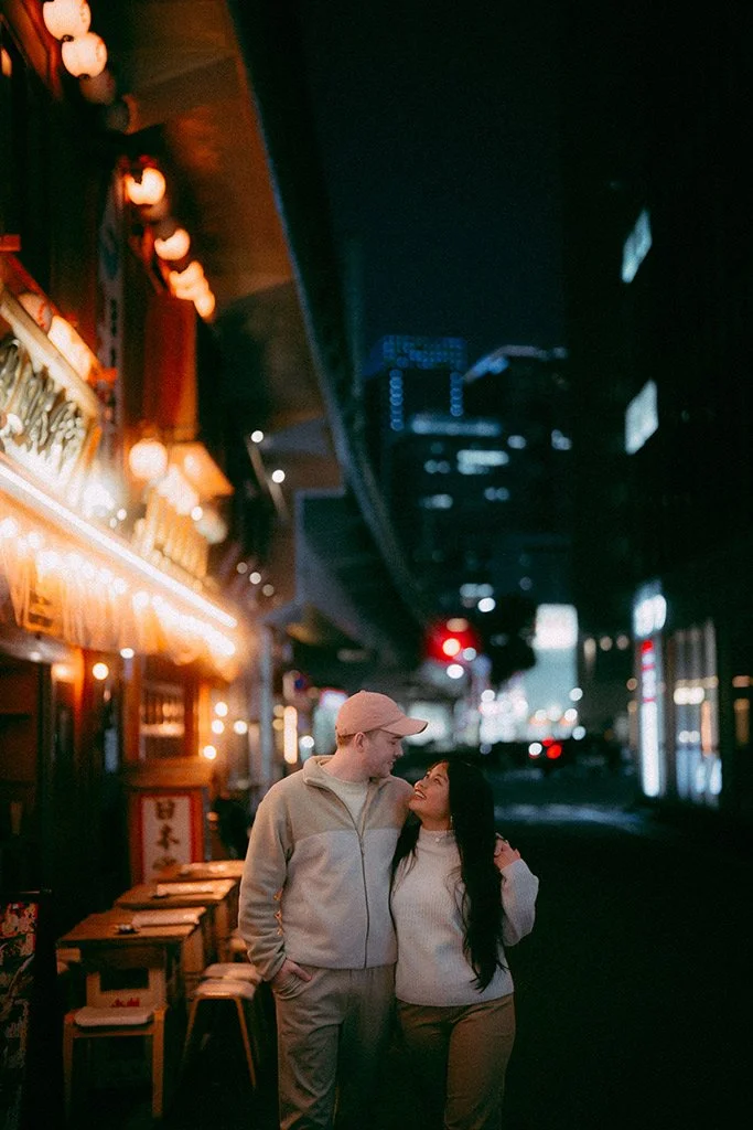 A couple stands close together on a city street at night, illuminated by warm restaurant lights, under a dark overpass with city lights and skyscrapers in the background.