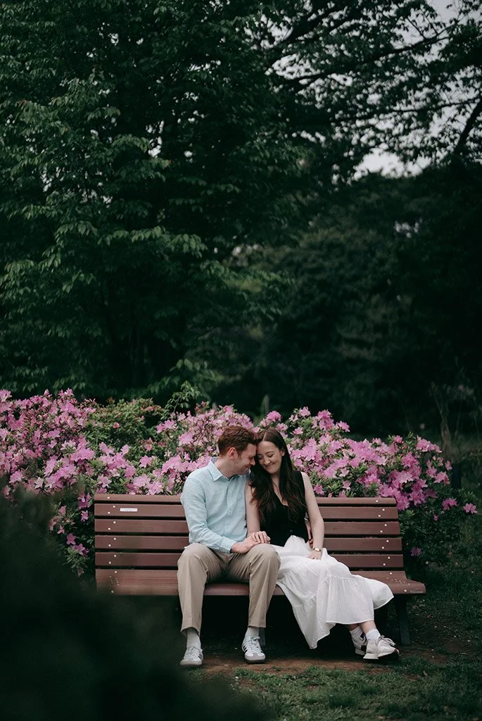 A young couple sits close together on a wooden park bench, surrounded by pink flowering bushes and green trees.