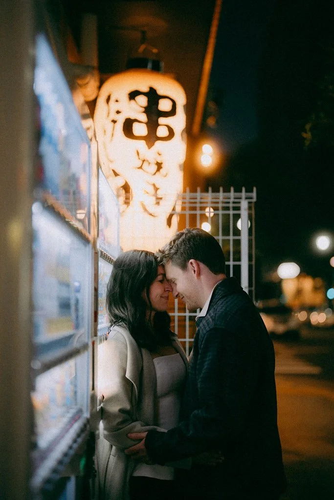 A couple stands close together at night, with foreheads touching and eyes closed, in front of a lit paper lantern with Japanese characters, outside a vending machine.