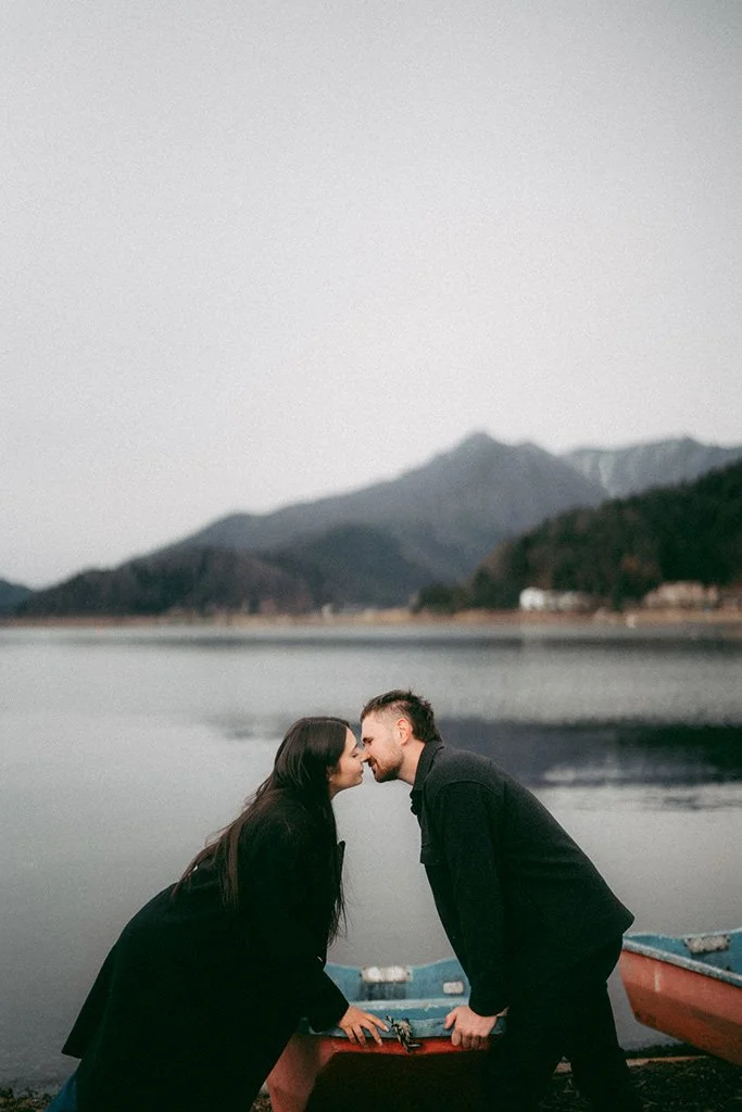 A couple leaning on a boat, nose to nose, with mountains and a lake in the background.