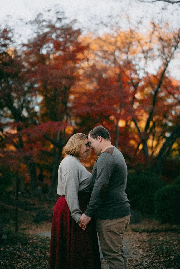 A couple holding hands and touching foreheads outdoors during autumn with red and orange trees in the background.