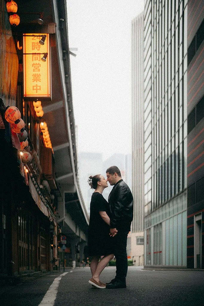 A couple standing close together with foreheads touching in an urban street surrounded by tall modern buildings and neon signs.