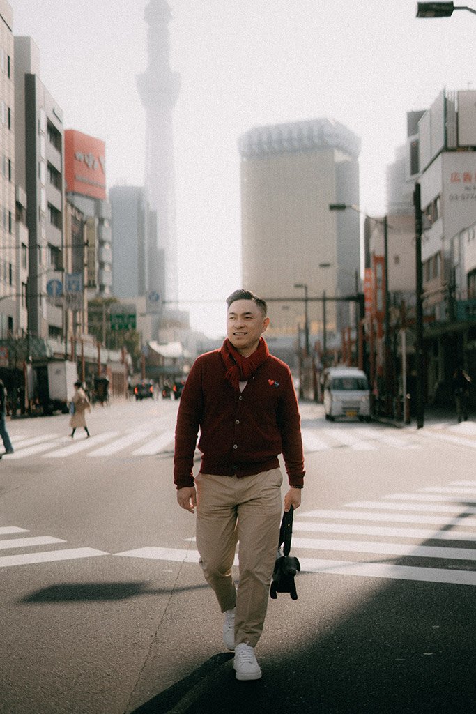 A man walking across a city street with tall buildings and a tower in the background, wearing a maroon sweater, beige pants, and white shoes.