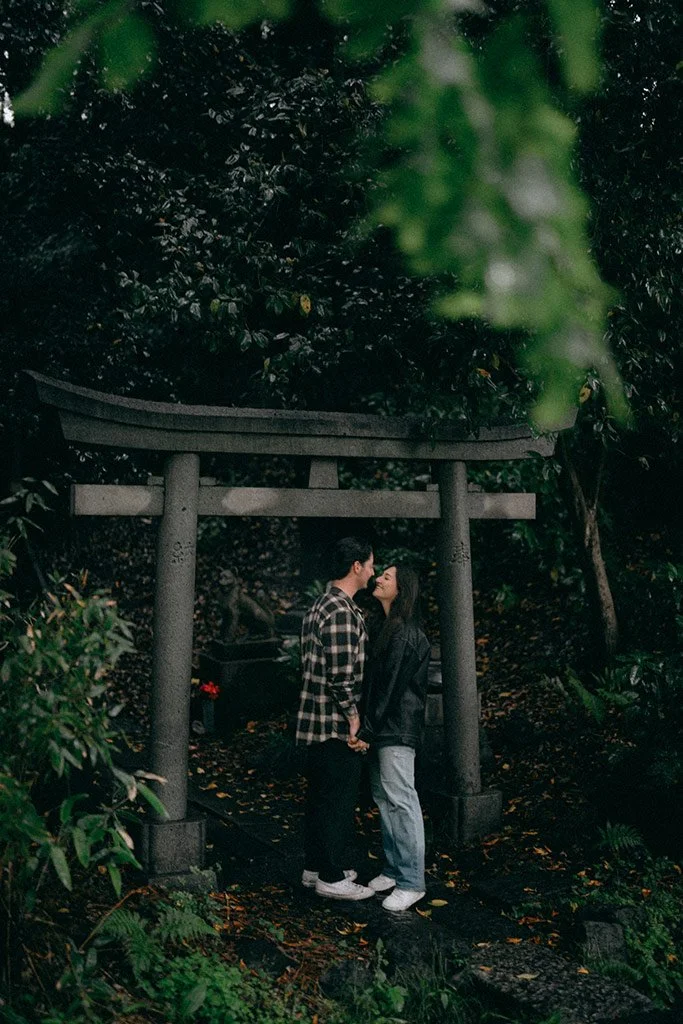 A couple standing close, about to kiss, beneath a Torii gate in a lush, green outdoor setting.