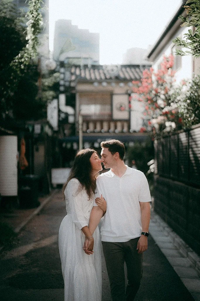 A couple is walking arm in arm on a street, leaning in close with their faces almost touching, smiling affectionately at each other. The woman is wearing a white dress, and the man is dressed in a white shirt and dark trousers. The background shows a