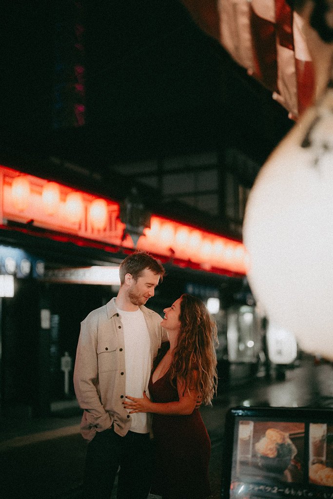 A couple stands close together on a city street at night, smiling and looking into each other's eyes, with red neon signs in the background.