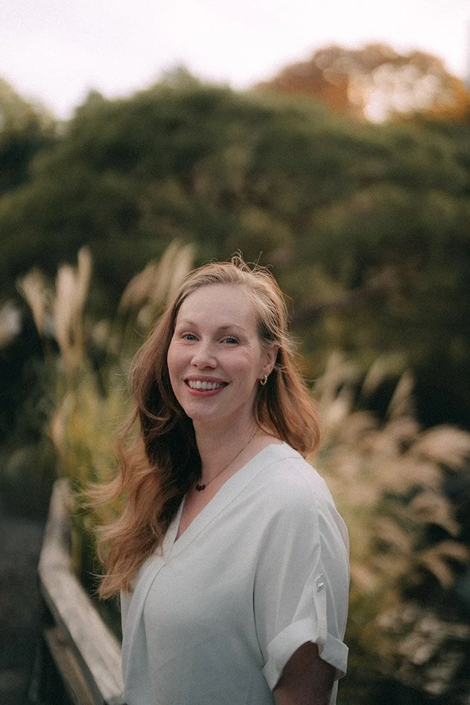 A young woman with long, wavy red hair smiling outdoors, wearing a white blouse with rolled-up sleeves, in a natural setting with greenery and blurred trees in the background.