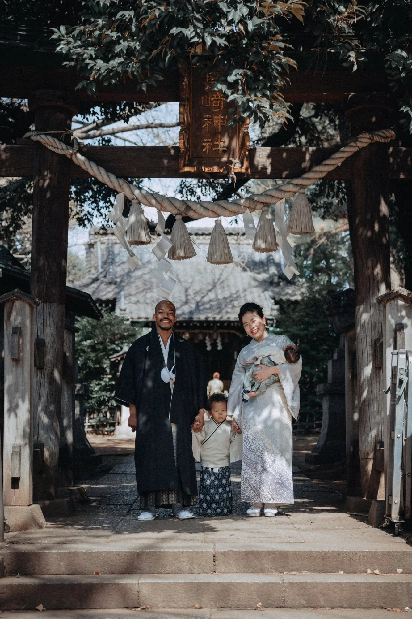 A Japanese family dressed in traditional kimono standing under a wooden torii gate at a Shinto shrine, with a man holding hands with a young boy and a woman holding a baby, smiling.
