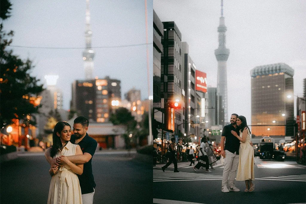 A couple standing in the middle of a city street at dusk, with a city skyline including a tall tower and buildings in the background. The woman is wearing a cream dress, and the man is in a black shirt and white pants. They are close together, smilin