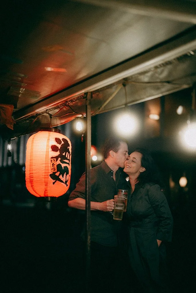 A man and woman sharing a kiss at an outdoor night market or festival, illuminated by a red Japanese paper lantern hanging from a stall.