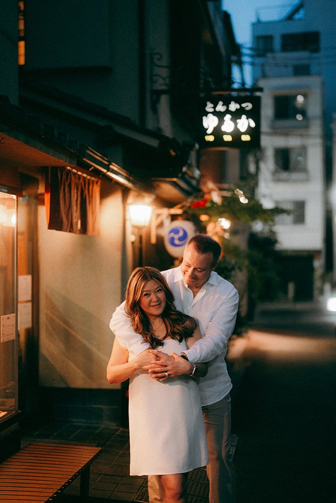 A man and a woman embrace on a city street at night, illuminated by warm lights from a nearby building, with Japanese signs on the building in the background.