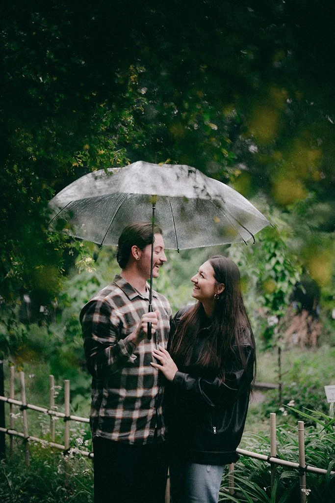 A couple smiling and looking at each other under a transparent umbrella in a lush green outdoor setting.