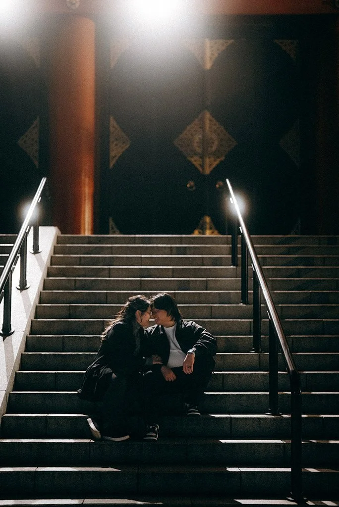 Two people sitting on stairs, leaning in for a kiss, with a dark background and railing on both sides.