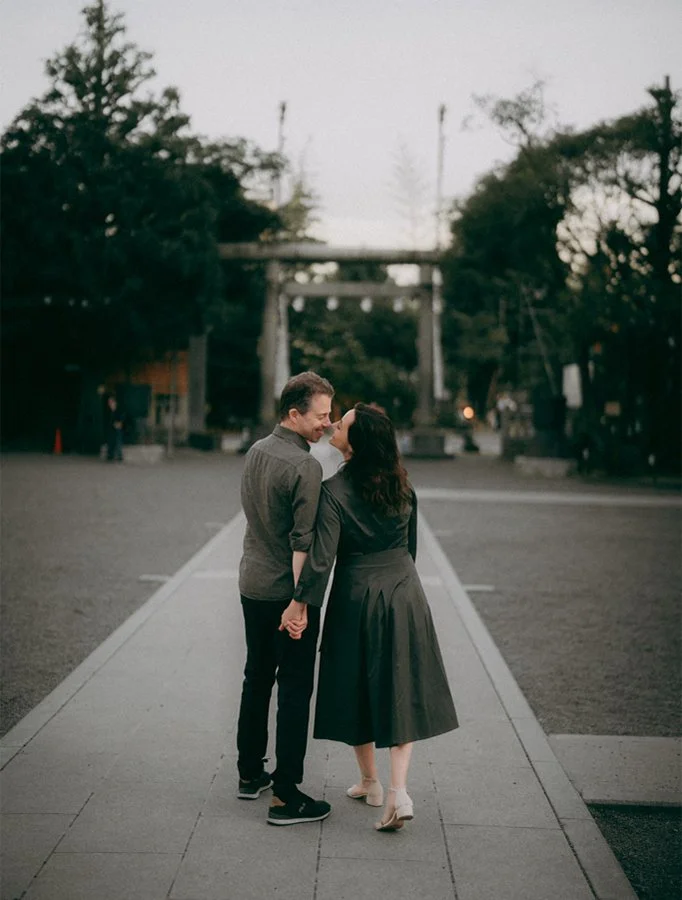 A couple holding hands and smiling at each other on a sidewalk during the evening or early night, with trees and a torii gate in the background.
