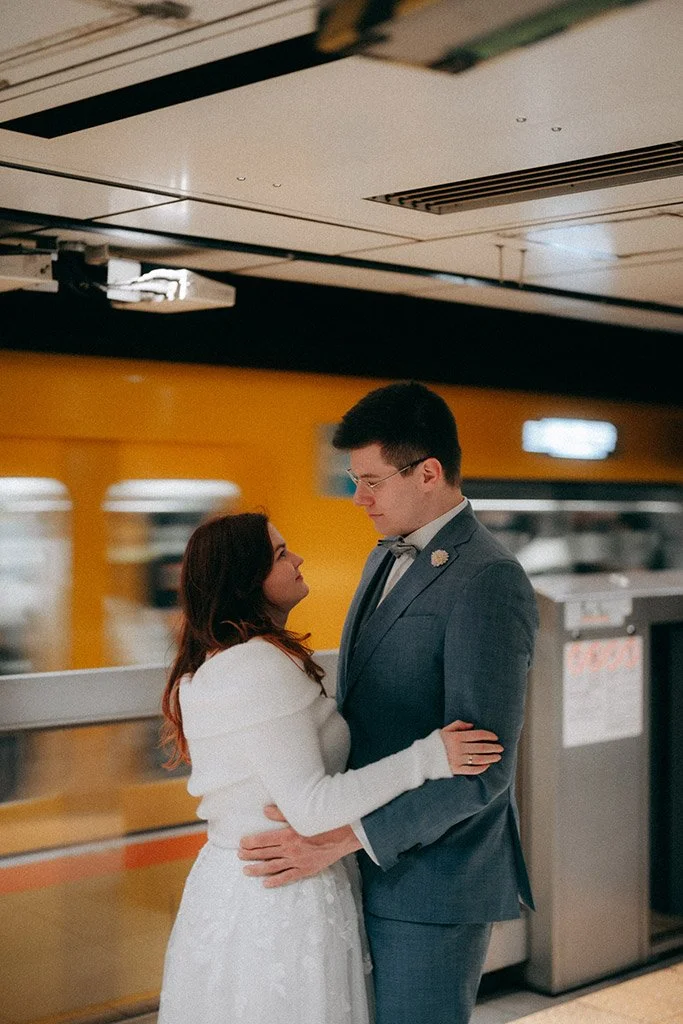A man and woman are dancing together indoors, dressed in formal attire, with the woman wearing a white dress and the man in a gray suit, in front of a yellow wall.
