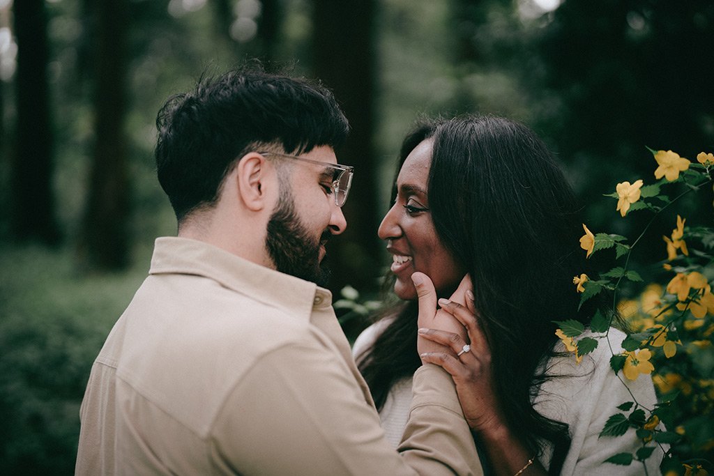 A couple stands close together outdoors, smiling and touching foreheads, with greenery and yellow flowers in the background.