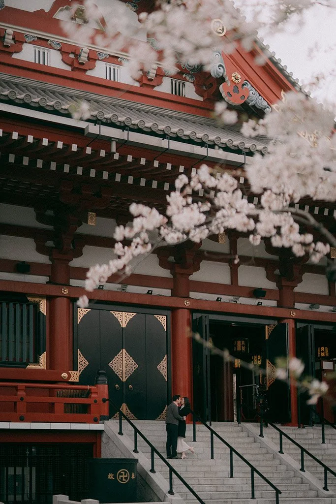 People standing and taking photos on the stairs outside traditional Japanese temple or shrine, with cherry blossoms in the foreground.