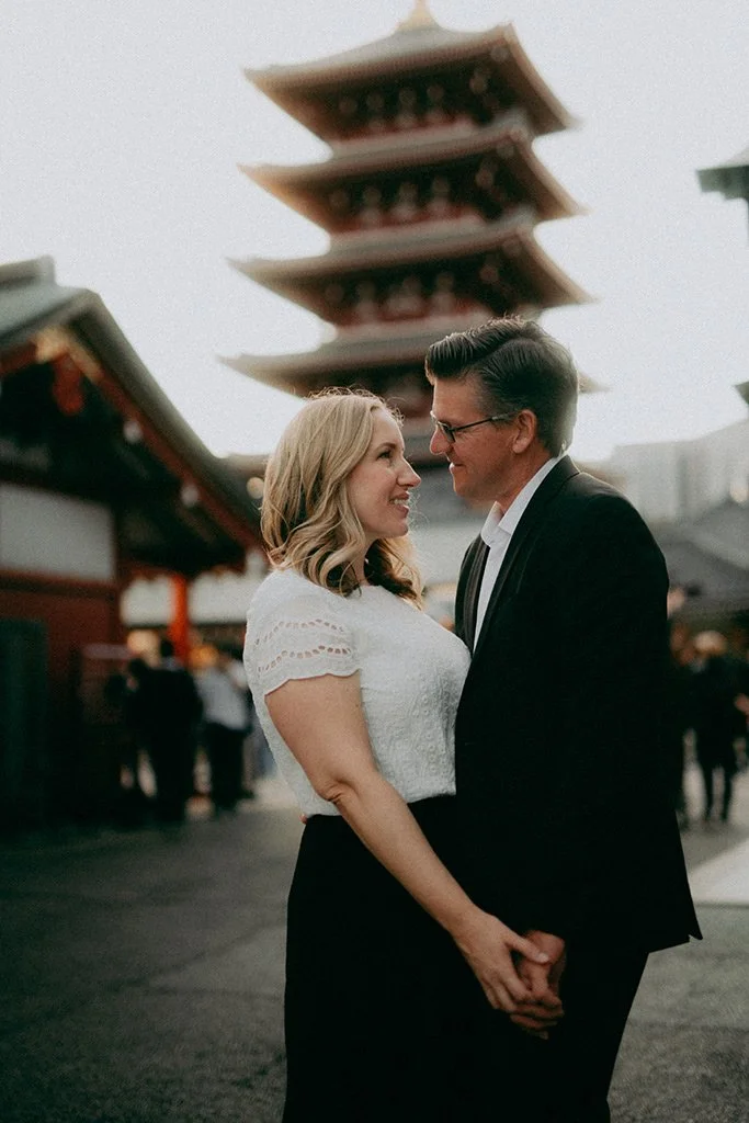 A couple stands close together, holding hands, in front of a traditional pagoda in an Asian temple or market setting, with other people in the background.