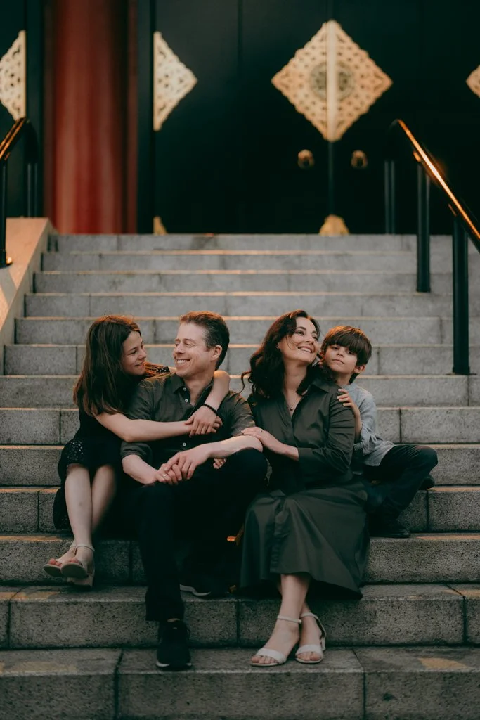 A family of four sitting on the stairs outside a building, smiling and enjoying each other's company.