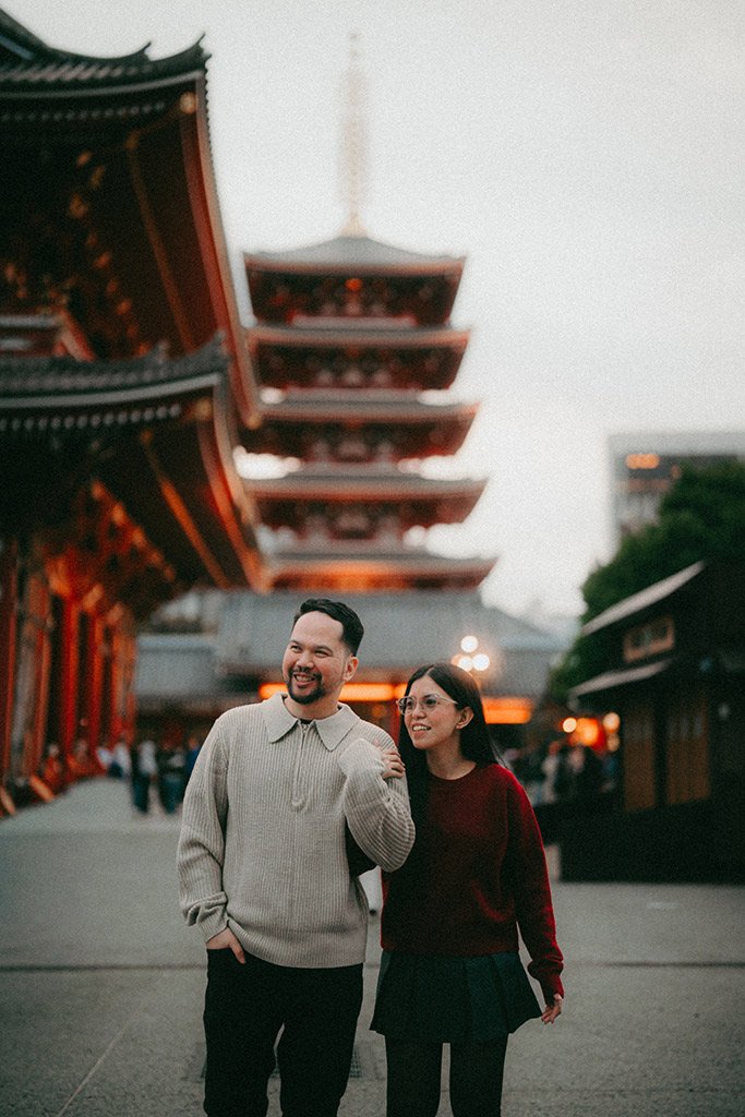 A smiling man and woman walking together at a traditional Japanese temple with pagoda rooftops in the background during evening.