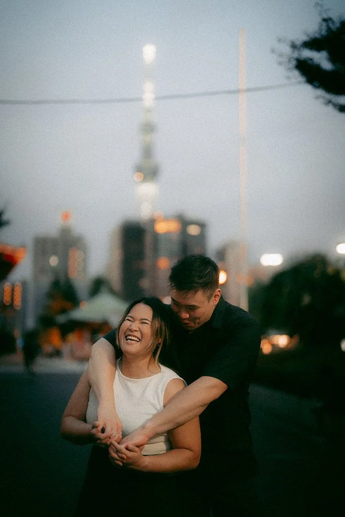 A smiling woman and a man are laughing and hugging each other outdoors in an urban setting at dusk, with a city skyline and tall building in the background.