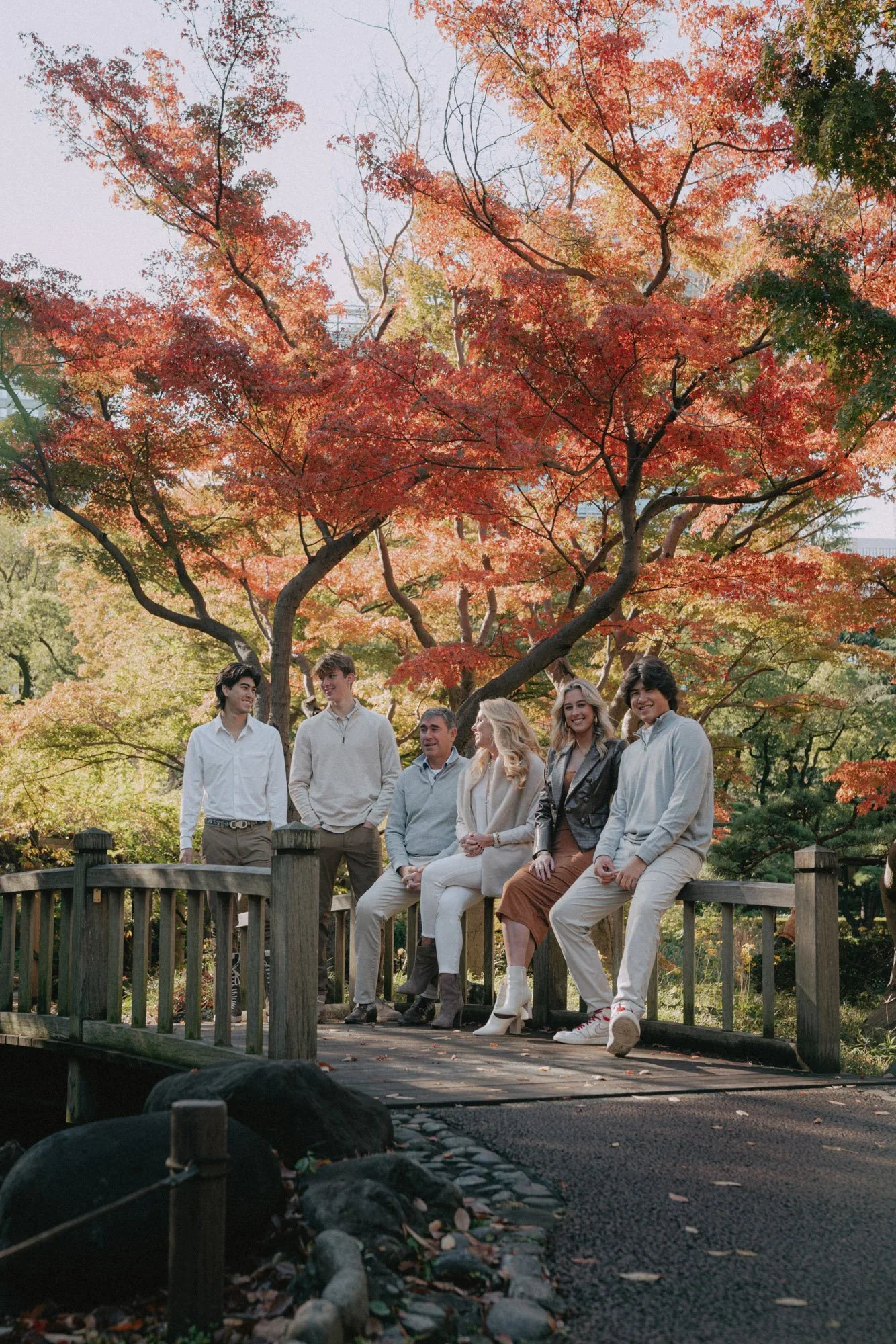 A group of six people are sitting and standing on a wooden bridge in a park during fall, with trees featuring orange, red, and green foliage in the background.