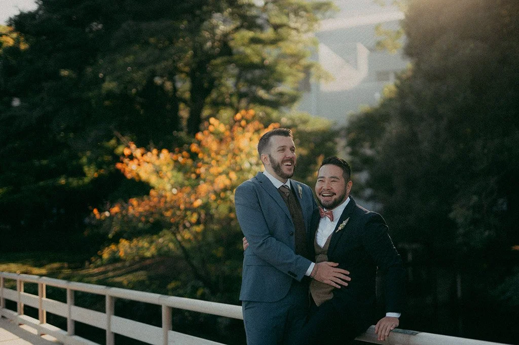 Two men in suits smiling and embracing each other on a bridge with autumn trees and a house in the background.