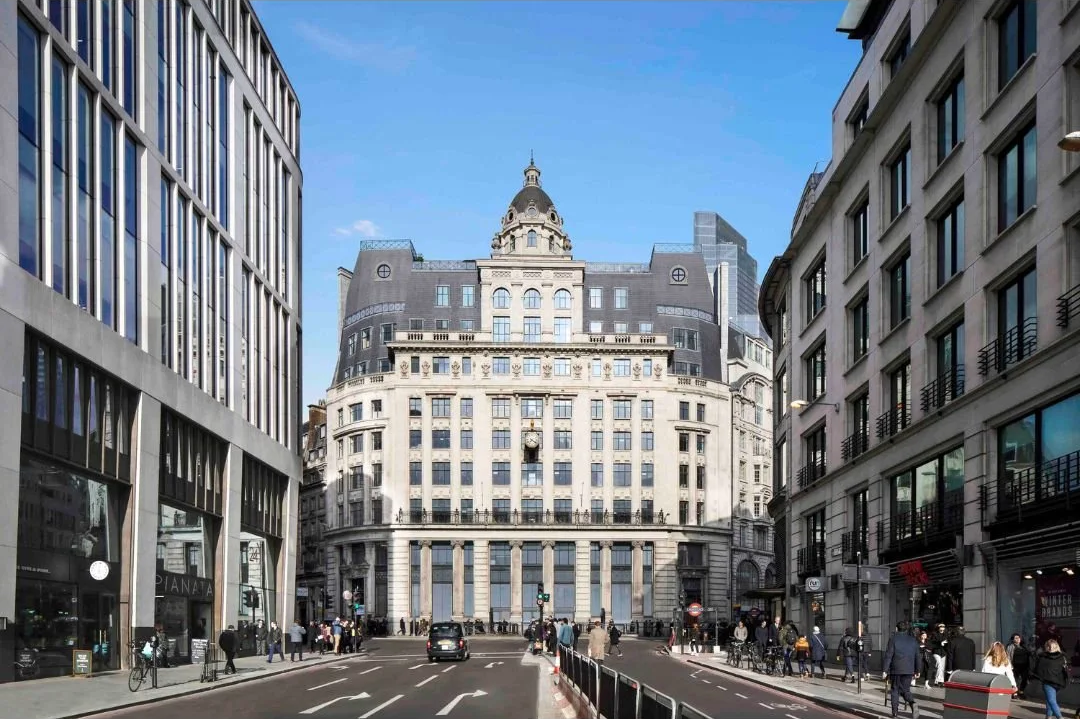 A city street surrounded by tall buildings on both sides, with a prominent historic multi-story building in the background, and pedestrians walking on the sidewalks.