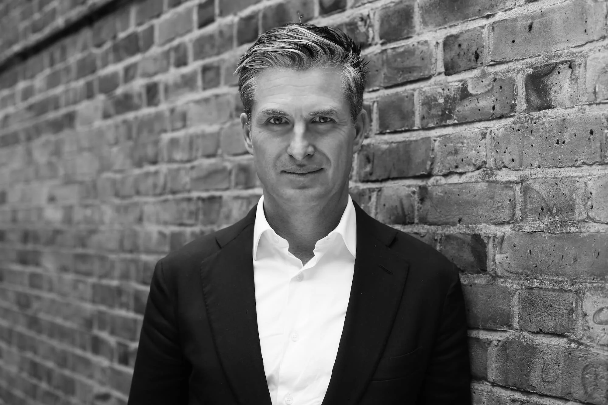 Oliver Corlette. Managing Partner. Black Mountain Partners, London, UK.  A black-and-white photograph of a man in a suit looking directly at the camera, standing against a brick wall.