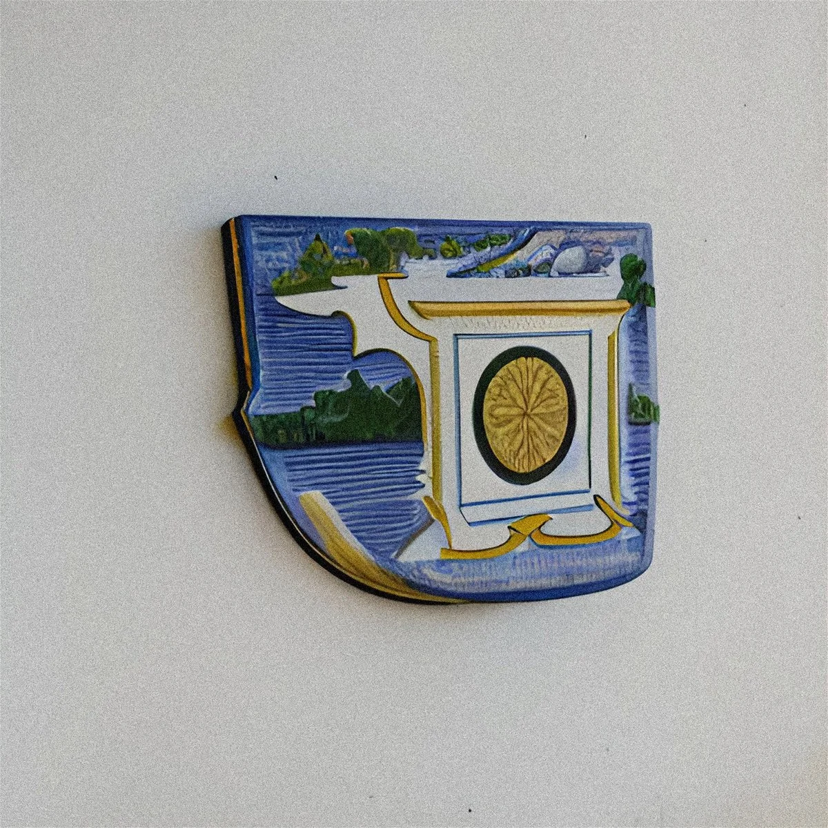 A colorful enamel badge featuring a detailed coat of arms with a blue background, a white and gold frame, and a central gold circle, on a plain light-colored surface.