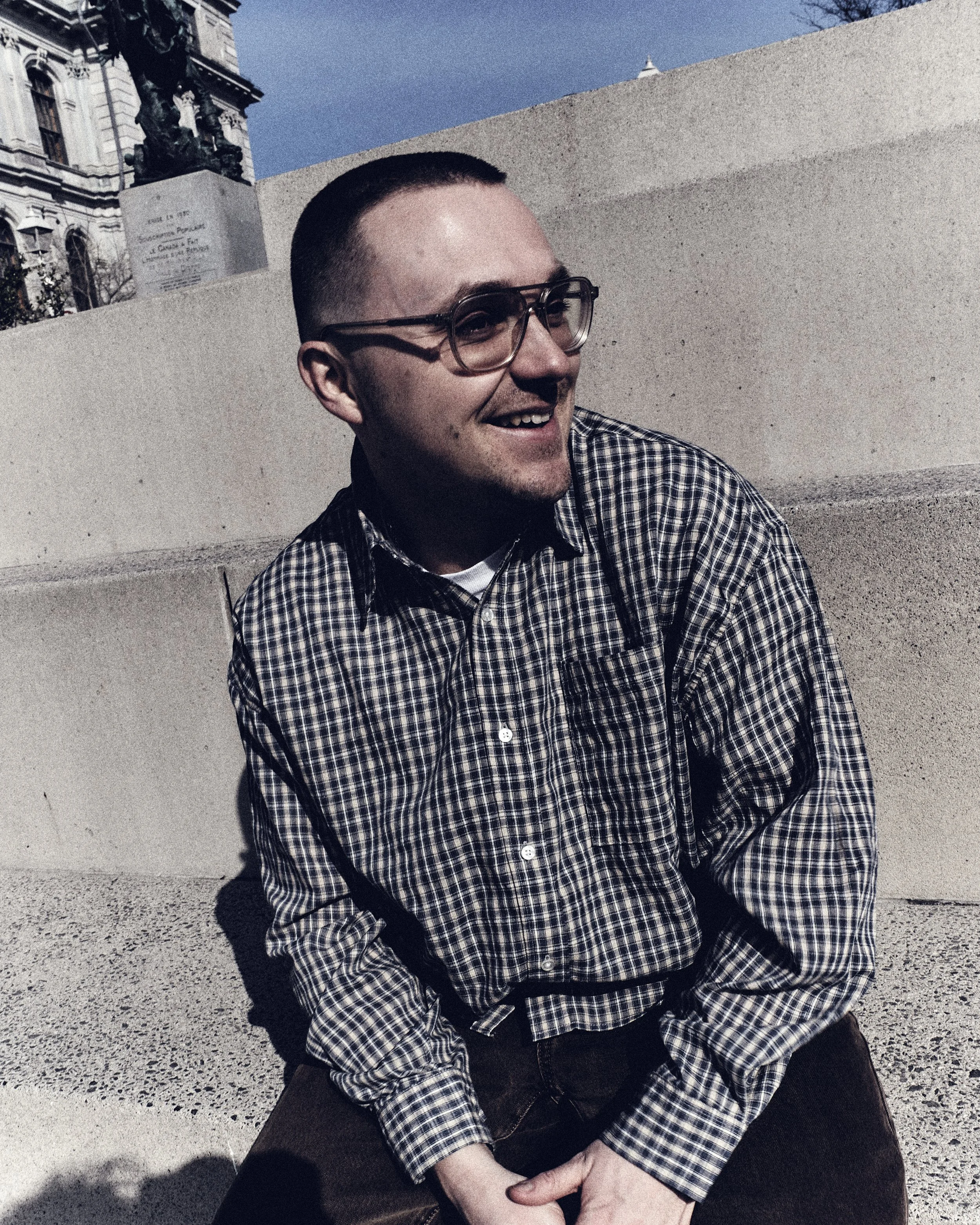 A young man with glasses, short hair, and a beard is sitting outdoors on a concrete surface, smiling. He is wearing a checkered shirt and black pants, and there is a building with ornate architecture in the background.