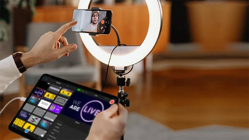 Person recording a live stream with a smartphone in front of a ring light, holding a tablet with stream control options, in an indoor setting.