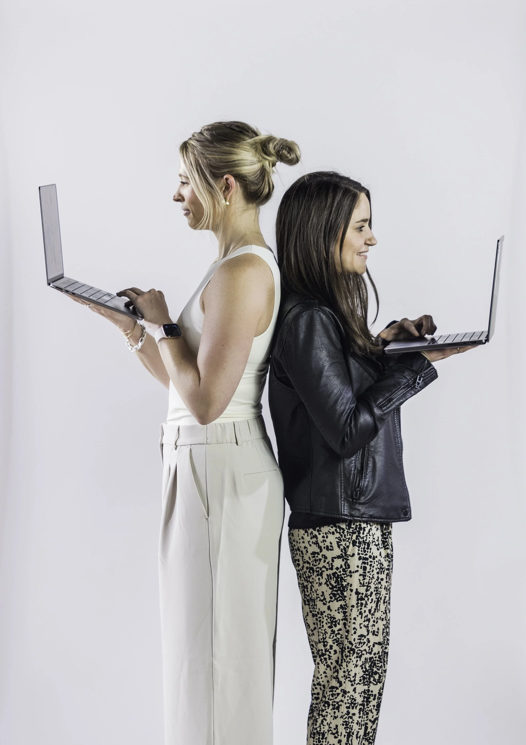 Two women stand back to back, each using a laptop, against a plain white background.