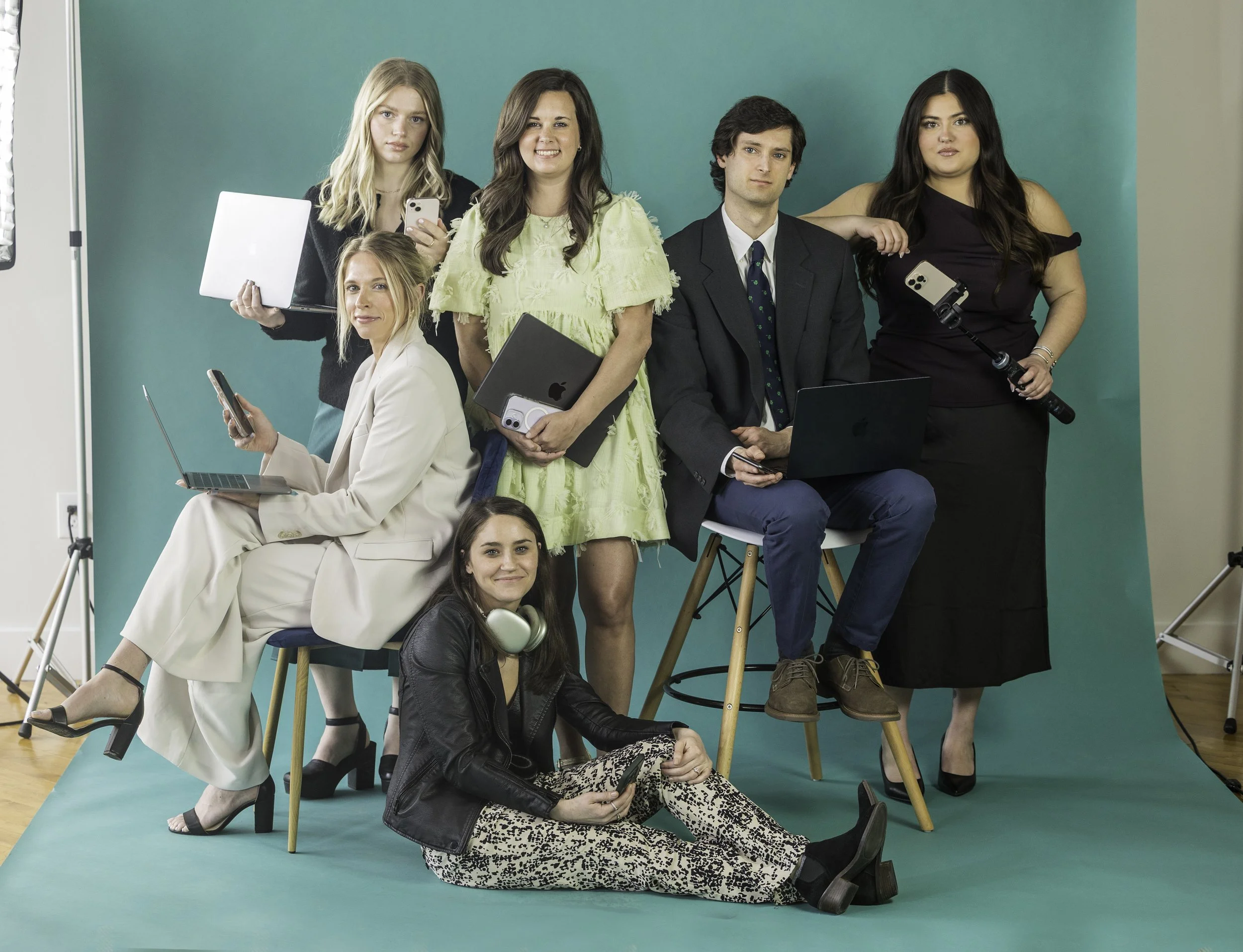 A group of seven diverse young adults, five women and two men, in a photoshoot studio with a teal backdrop, holding various electronic devices and posing confidently.