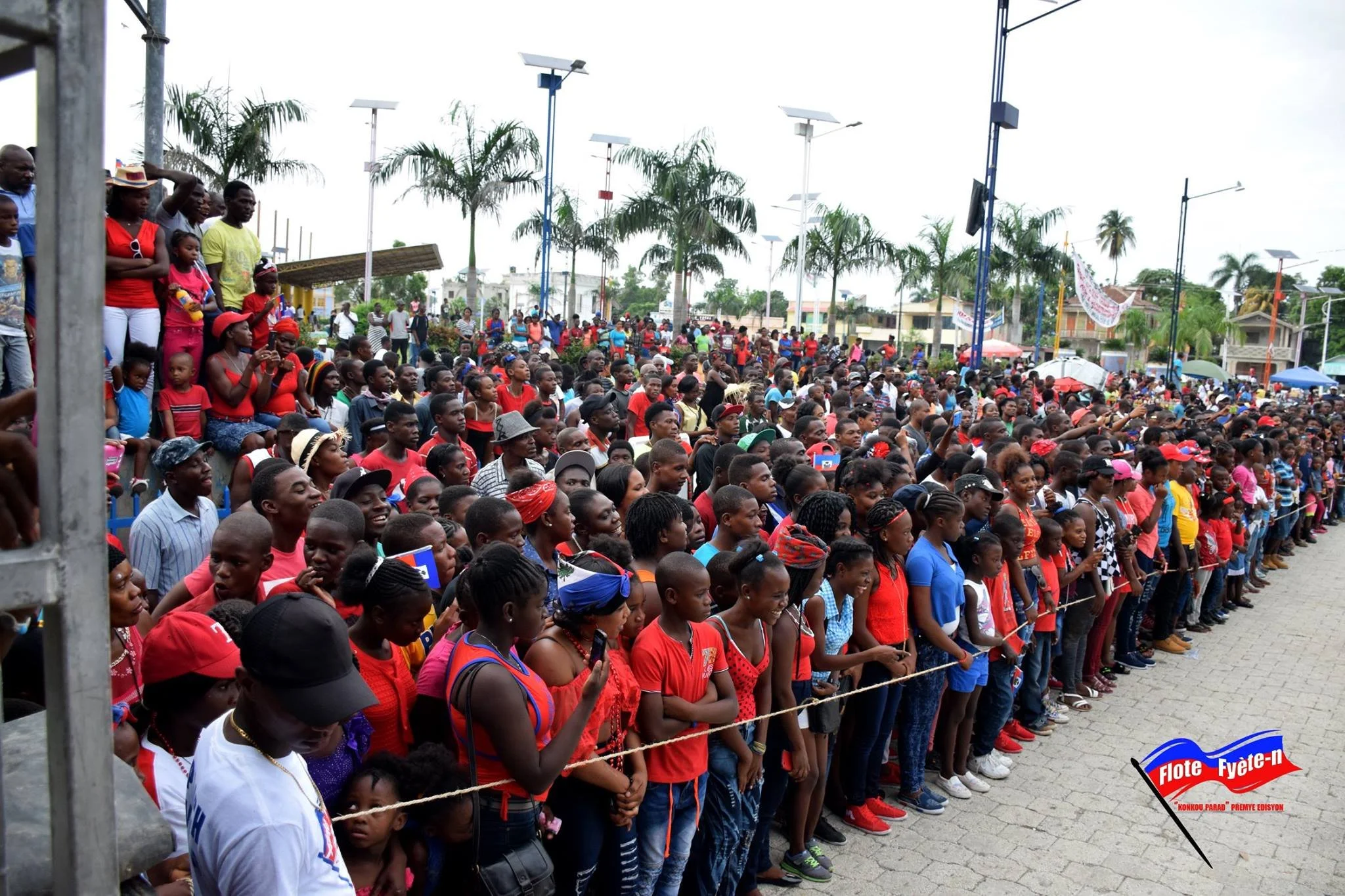 Une grande foule de personnes, principalement des enfants, assistent à un événement public dans une zone en plein air avec des palmiers et des lampadaires. Beaucoup portent des vêtements rouges. Il y a un drapeau avec le texte 'Flote Fête-n' dans le 