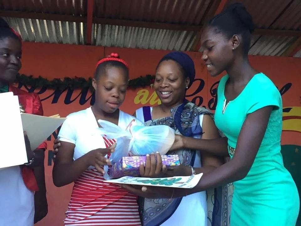 Cinq femmes et une fille échangent un cadeau lors d'un événement, toutes souriantes, contre un fond orange décoré vert.