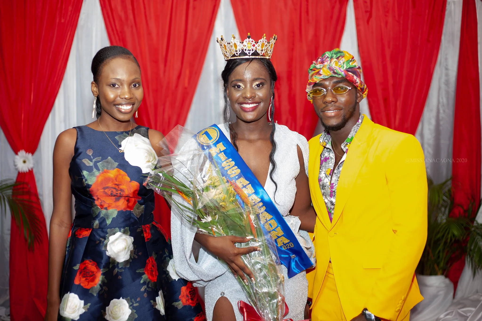 Une femme portant une couronne et un sash de Miss annonce un concours, entourée de deux personnes habillées de façon colorée, avec un fond de rideaux rouges.