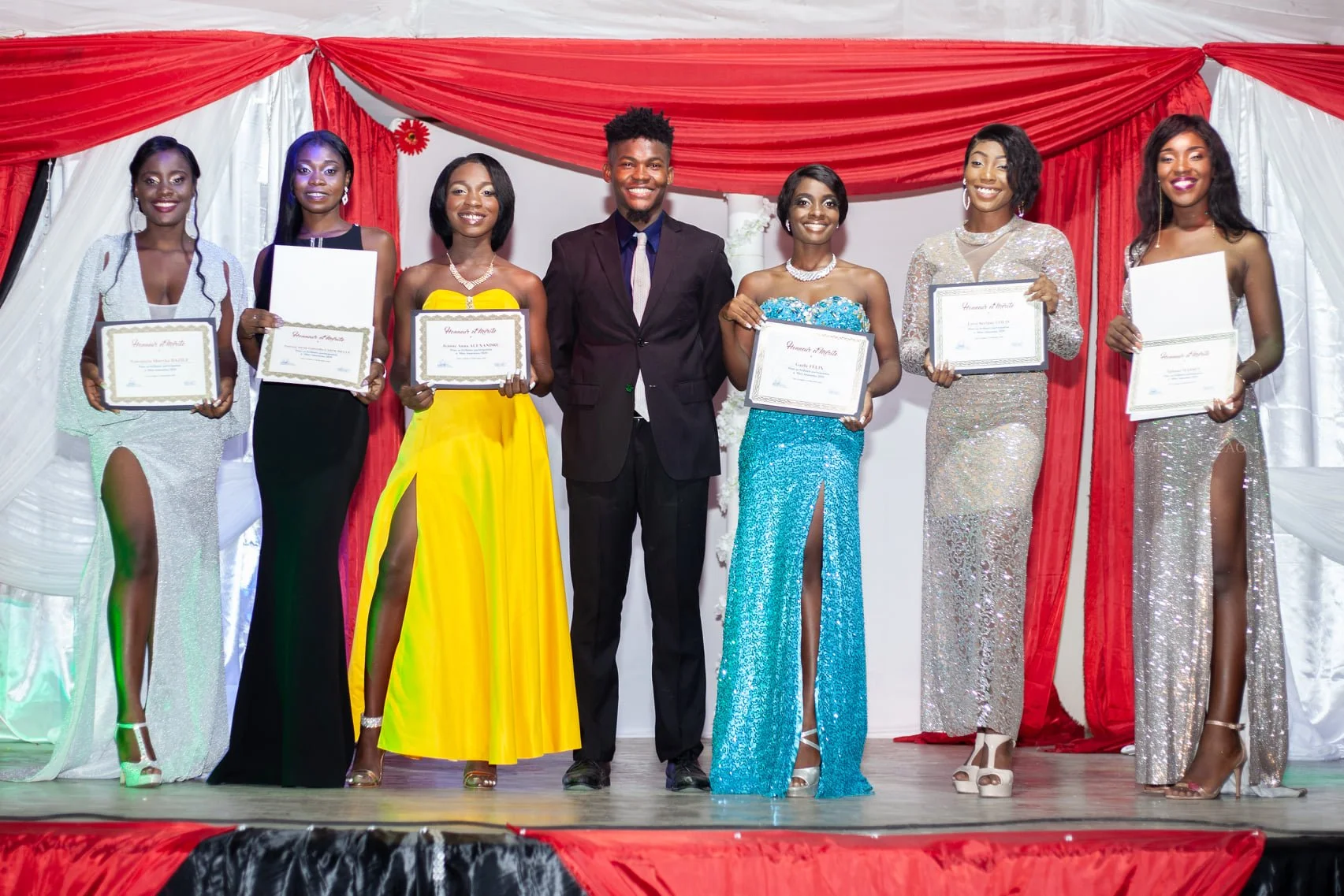 Groupe de sept personnes en robes de soirée et costume, tenant des certificats, sur scène avec un fond décoré de tissu rouge et blanc, lors d'une cérémonie de remise de prix ou diplômes.