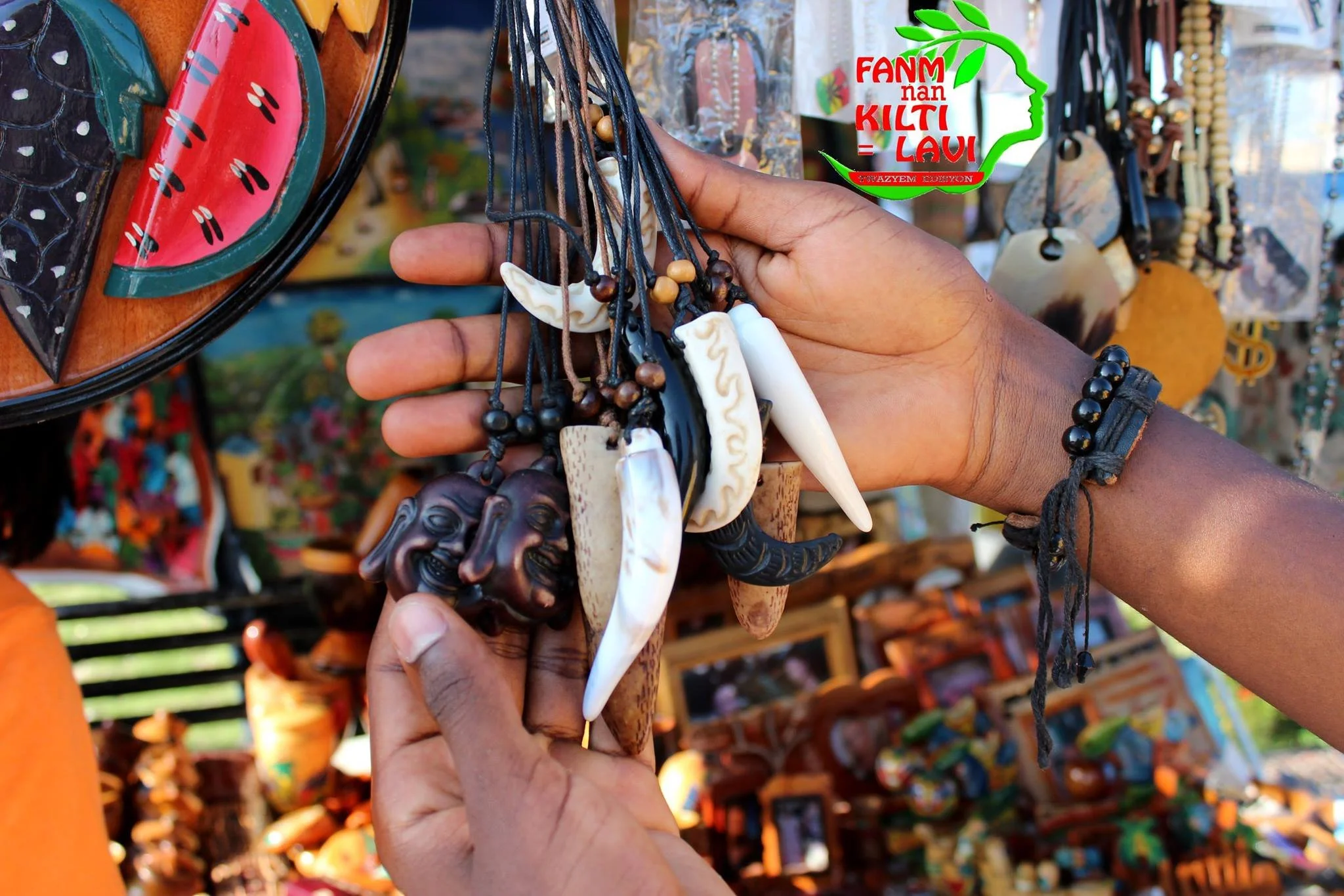 Main dans une main tenant divers colliers en ivoire ou matériaux similaires, avec des objets tribaux ou artisanaux, devant un stand de souvenirs ou de l'artisanat africain, avec des bijoux et des objets en bois ou en pierre en arrière-plan.