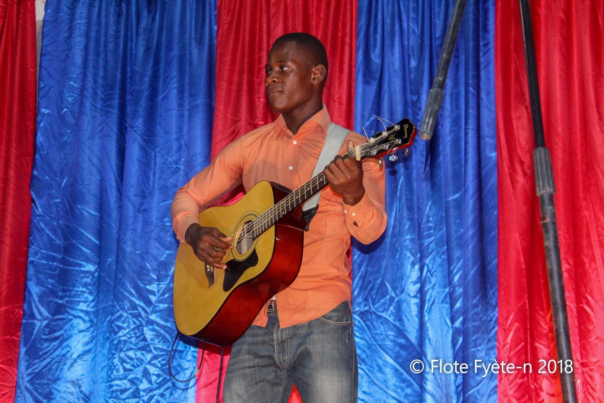 Un jeune homme joue de la guitare acoustique sur scène, en arrière-plan un rideau bleu et rouge.