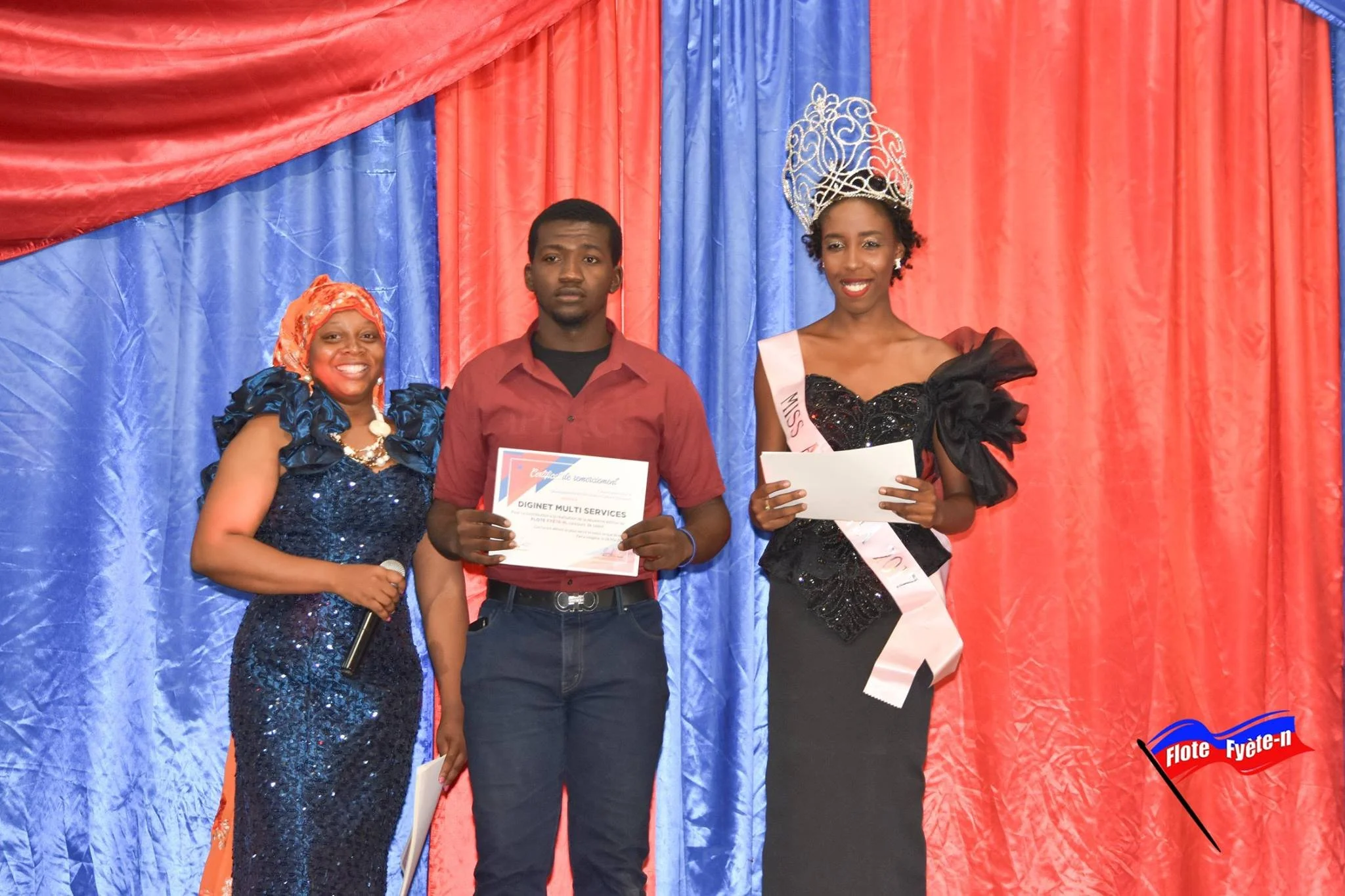 Une cérémonie de remise de prix ou de reconnaissance avec une femme portant une couronne et une écharpe de Miss, un homme recevant un certificat, et deux autres femmes portant des tenues élégantes, devant un fond en draps rouge et bleu.
