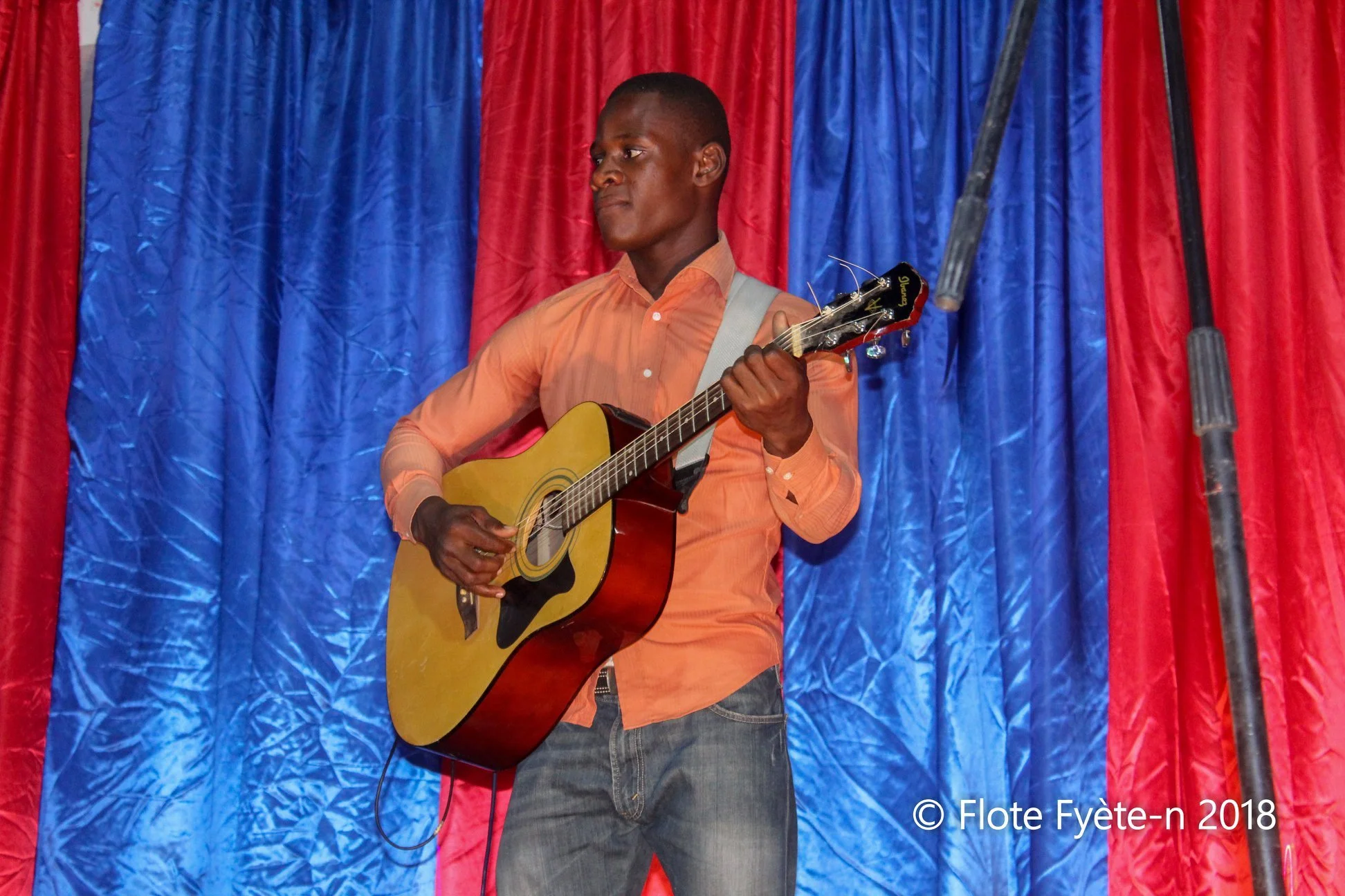 Un homme jouant de la guitare sur scène avec un décor de rideaux rouges et bleus en arrière-plan.
