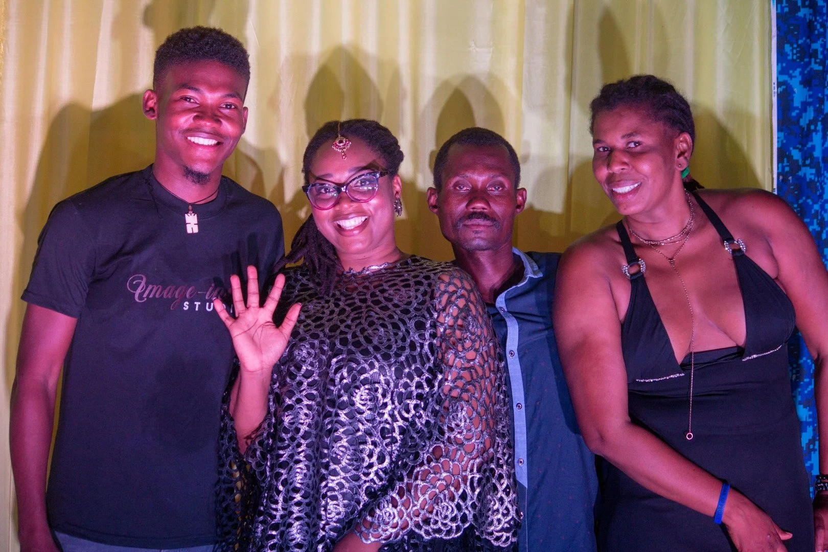 Quatre personnes posant pour une photo en intérieur avec un fond jaune, deux femmes et deux hommes, souriants, certains faisant un signe de la main.