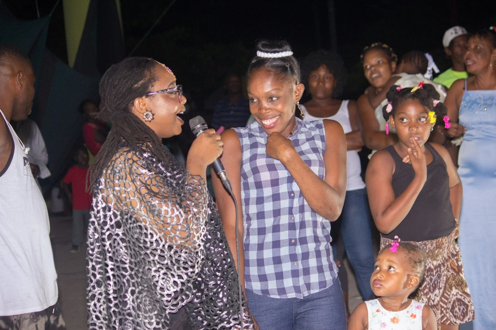 Groupe de personnes lors d'un événement social, une femme avec un micro sourit à une autre femme, entourés d'enfants et d'adultes.