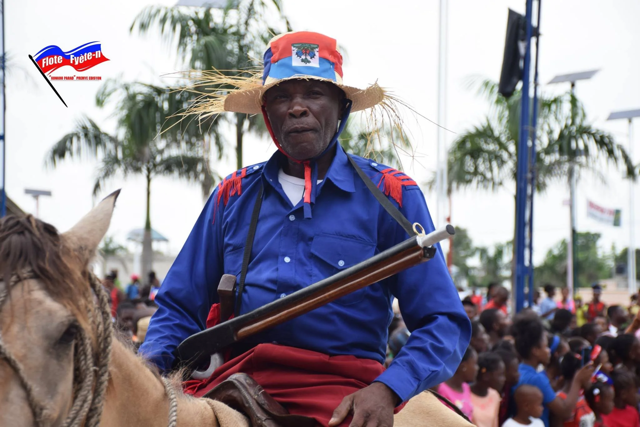 Un homme en costume traditionnel haïtien, portant un chapeau décoré du drapeau d'Haïti, à cheval lors d'une fête ou parade, avec un groupe de personnes en arrière-plan.