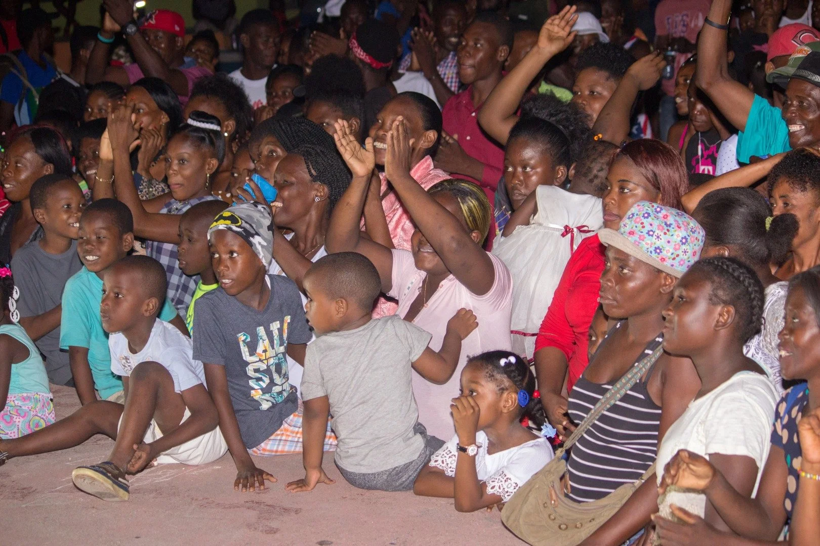 Une foule de personnes, dont de nombreux enfants, assises et debout, assiste probablement à un événement ou spectacle, manifestant de la joie et de l'enthousiasme.
