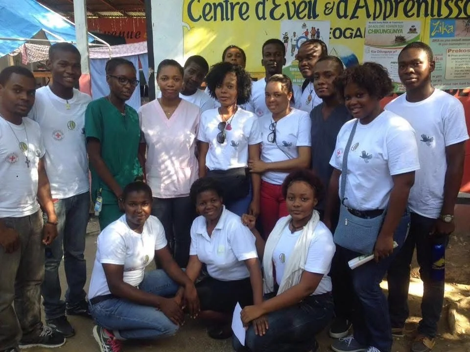 Groupe de personnes posant devant un panneau indiquant un centre d'éveil et d'apprentissage, avec plusieurs individus portant des T-shirts blancs ou verts, certains avec des badge ou du matériel médical, à l'extérieur, lors d'un événement ou activité